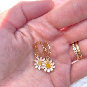 Peut inclure: Une paire de boucles d'oreilles cr&eacute;oles dor&eacute;es avec un petit charme de marguerite blanche suspendu &agrave; chaque cr&eacute;ole. Les marguerites ont des centres jaunes.