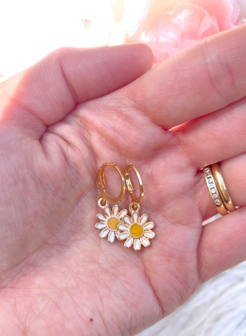 Peut inclure: Une paire de boucles d'oreilles cr&eacute;oles dor&eacute;es avec un petit charme de marguerite blanche suspendu &agrave; chaque cr&eacute;ole. Les marguerites ont des centres jaunes.