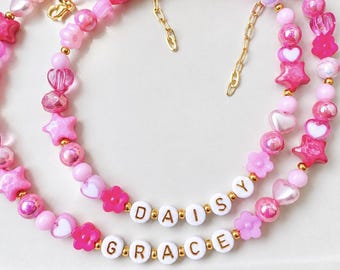 May include: Two layered necklaces featuring pink and white beads in various shapes including stars, hearts, and flowers. The necklaces include white round beads with gold letters spelling DAISY and GRACE, accented with small gold-tone metal beads.