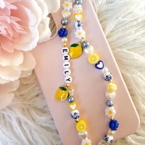 May include: A decorative phone charm featuring a mix of beads: white daisies, yellow hearts, blue roses, and lemon charms. The charm spells "EMILY" with white letter beads. The charm is displayed on a pink phone case.