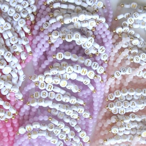 May include: A close-up of several beaded bracelets with the word "breathe" spelled out in white beads with gold lettering. The bracelets are in shades of pink, lavender, and light pink.