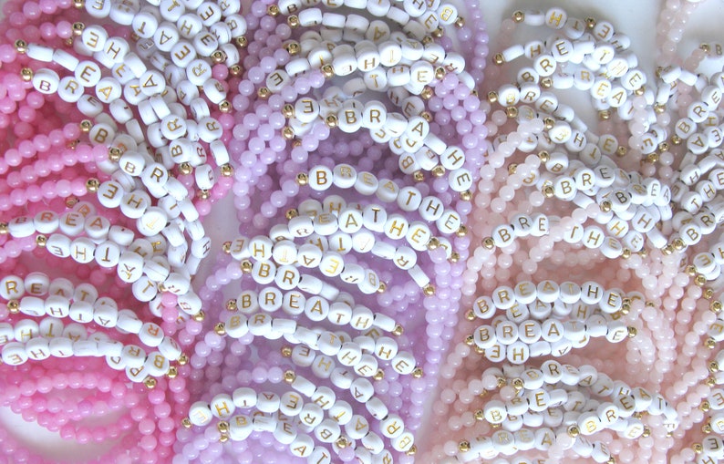 May include: A close-up of several beaded bracelets with the word "breathe" spelled out in white beads with gold lettering. The bracelets are in shades of pink, lavender, and light pink.