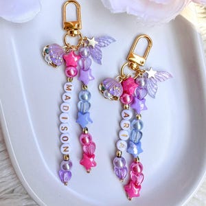 May include: Two personalised keychains with gold-coloured clasps. Each keychain features a beaded design with pink, purple, and blue beads, along with a name spelled out in white letter beads. Decorative charms include stars, shells, and wings.