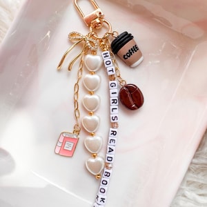 May include: Gold-coloured keychain with various charms. Charms include a coffee cup, coffee bean, pink book, and white heart beads. The keychain also has a strand of letter beads that spell out "HOT GIRLS READ BOOKS".