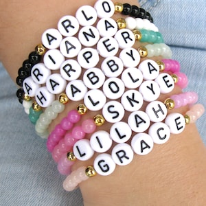 May include: A stack of colorful beaded bracelets with white letter beads spelling out the names "Arlo", "Ariana", "Harper", "Abby", "Lola", "Skye", "Lila", and "Grace".