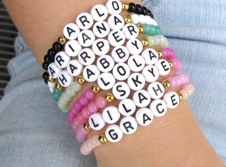 May include: A stack of colorful beaded bracelets with white letter beads spelling out the names "Arlo", "Ariana", "Harper", "Abby", "Lola", "Skye", "Lila", and "Grace".
