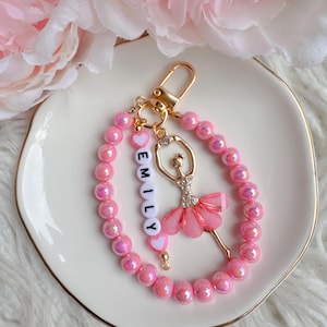 May include: Pink and gold ballerina keychain with a pink pearl chain and the name "EMILY" spelled out in white beads.