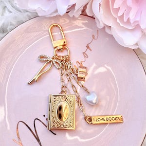 May include: Gold-tone keychain featuring a book charm, a heart, and a bow. The keychain includes a charm with the text "I LOVE BOOKS". The background is a pink plate with a floral arrangement.