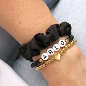 May include: A black scrunchie with a gold beaded bracelet and a white beaded bracelet with the name "ARLO" spelled out in white letters. A gold heart charm hangs from the bracelet.