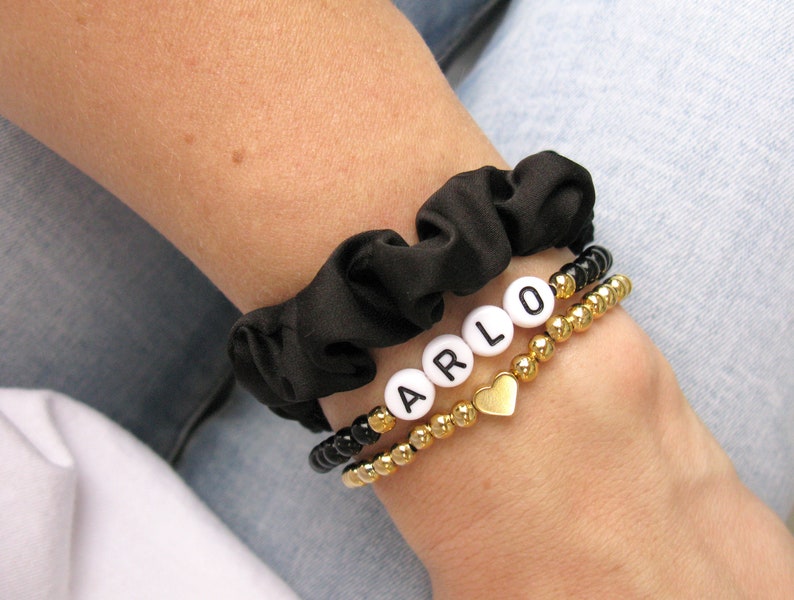 May include: A black scrunchie with a gold beaded bracelet and a white beaded bracelet with the name "ARLO" spelled out in white letters. A gold heart charm hangs from the bracelet.