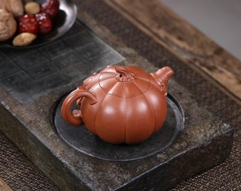 May include: A red clay teapot shaped like a pumpkin with a lid and a handle. The teapot is sitting on a dark gray stone surface.