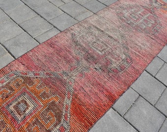 Puede incluir: Una alfombra vintage roja y naranja con un patrón geométrico. La alfombra está colocada sobre una superficie de hormigón gris.