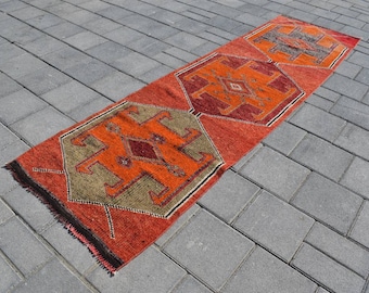 Könnte beinhalten: Ein langer, schmaler Teppich mit traditionellem türkischem Design. Der Teppich ist rot mit orangefarbenen, braunen und schwarzen geometrischen Mustern. Der Teppich liegt auf einer grauen Betonfläche.