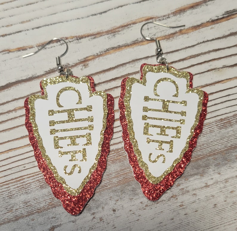 May include: A pair of red and gold glitter arrowhead earrings with the word "Chiefs" written in gold glitter on each earring.