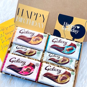 May include: A gift box filled with six Galaxy chocolate bars. The box is lined with brown paper and includes two yellow cards with the text "Happy Birthday".