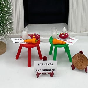 May include: A miniature Christmas scene with a white fireplace and black top, a small Christmas tree, and a gray cat figurine in a sweater. Two small tables hold treats and a sign that reads "FOR SANTA AND RUDOLPH".