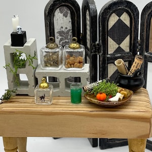 Puede incluir: Miniatura con temática de Halloween que presenta una mesa de madera con varios objetos. Incluye frascos de vidrio con tapas doradas, un pequeño caldero negro, una calabaza en miniatura y una bebida verde. Una calavera y una vela están sobre un bloque de hormigón.