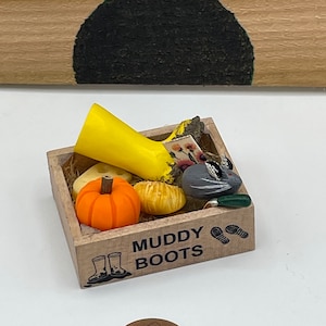 Puede incluir: Una pequeña caja de madera con las palabras "Muddy Boots" impresas en la parte delantera. La caja está llena de artículos en miniatura, incluyendo una bota de lluvia amarilla, una calabaza naranja y un pájaro gris.