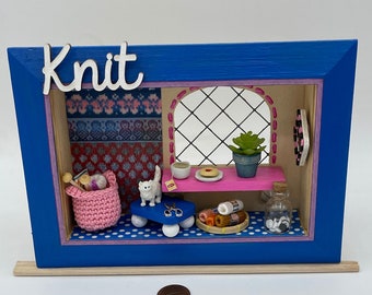 Könnte beinhalten: Ein blauer Holzrahmen mit dem Wort "Knit" auf der Vorderseite. Im Rahmen befindet sich eine Miniatur-Szene eines Strickplatzes mit Garn, Nadeln, einer Katze und einer Tasse Tee.