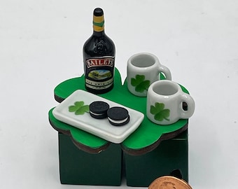 May include: A miniature green shamrock-shaped table with two white mugs, a bottle of Bailey's Irish Cream, two chocolate cookies, and a small white plate. The table is on a white surface with a penny on a stand.
