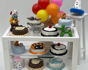 May include: Miniature display of birthday cakes, balloons, and a clown figurine. The cakes feature various designs, including a ladybug, monkey, and bear. A party hat and a green balloon dog are also present. A penny is displayed in the foreground.