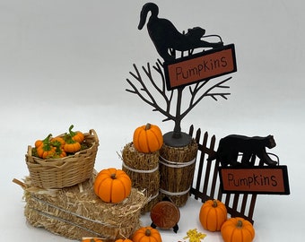 May include: A miniature autumn display featuring orange pumpkins, a woven basket, straw bales, and two black metal signs with the text Pumpkins. Includes a black cat silhouette, a wooden fence, a copper penny for scale, and scattered colourful fall leaves.