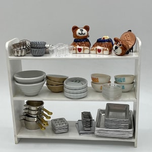 Puede incluir: Una estantería blanca de tres niveles, llena de diversos utensilios de cocina, incluyendo cuencos de cerámica, moldes para hornear de metal y tazas medidoras. También hay dos pequeñas figuritas de osos de peluche de cerámica en la repisa superior.