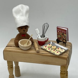 Pode incluir: Um urso de peluche marrom em miniatura vestindo um chapéu de chef branco está assando biscoitos em uma mesa de madeira. O urso está segurando uma tigela de madeira e uma colher de madeira. Há um rolo de massa, uma caneca vermelha e branca, uma espátula, um batedor e uma assadeira com biscoitos na mesa. Um livro de receitas em miniatura intitulado "Betty Crocker's Cooky Book" está em um suporte ao lado da assadeira. Há um centavo no chão na frente da mesa.