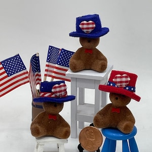 Puede incluir: Tres osos de peluche marrones que llevan sombreros patrióticos rojo, blanco y azul. Los osos están sentados en taburetes blancos y azules. Los osos están rodeados de banderas americanas.