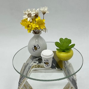 May include: A miniature hexagonal table with a glass top, decorated with a bee motif. The table features a white vase with yellow and white flowers, a small white cup with a bee design, and a yellow planter with a green succulent.