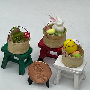 May include: Three miniature wooden stools with baskets filled with Easter-themed figurines. The stools are painted green, red, and white. The baskets contain a frog, a bunny, and a chick.