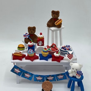 May include: A miniature scene with two brown teddy bears, one white teddy bear, and a variety of miniature food items, including cupcakes, hot dogs, watermelon, and a hamburger. The scene is set on a white table with a blue and white bunting and a penny on a stand.