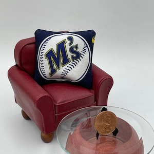 Könnte beinhalten: Ein roter Miniatur-Sessel mit einem blauen und weißen Baseball-Kissen mit dem Text "M's". Ein kleiner Glastisch mit roter Basis steht vor dem Sessel.