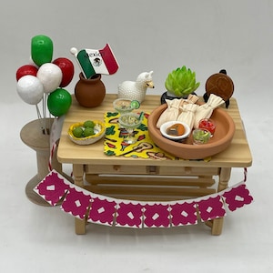 May include: A miniature wooden table set with a Mexican fiesta theme. The table is decorated with a green and red flag that says "Mexico", a llama figurine, a succulent plant, and a string of pink paper flags. There are also miniature tamales, a margarita glass, and a bowl of green olives.