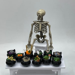 May include: A miniature skeleton sits on a white pedestal table with eight black cauldrons filled with various Halloween-themed items, including a snake, a pumpkin, a nest, mushrooms, and a cat. A penny is in the foreground for scale.