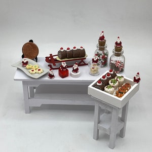 May include: A miniature Christmas-themed bakery display with a white table and a white stand. The table is decorated with a variety of miniature Christmas treats, including cookies, cupcakes, and a chocolate Yule log. There are also miniature Santa Claus figurines and jars filled with candy.