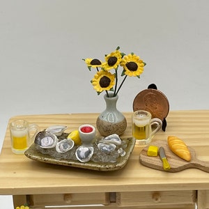 May include: A miniature table setting with a plate of oysters on ice, two glasses of beer, a small bowl of red sauce, a lemon wedge, a butter knife, a loaf of bread, and a vase of sunflowers.