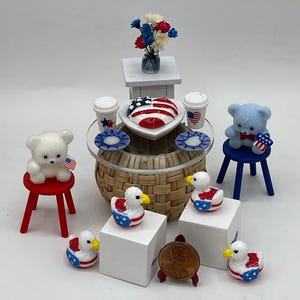 May include: Miniature patriotic-themed decor featuring a small table with a heart-shaped dish, coffee cups, and plates. Includes teddy bears, ducks, and a penny, all decorated with American flag motifs. The scene is set on a white surface.