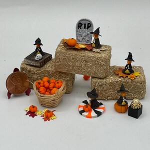 Może przedstawiać: Miniaturowe dekoracje na Halloween, w tym bele siana, dynie, figurki czarownic i nagrobek z napisem "RIP". W zestawie znajduje się również kosz pomarańczowych dyń i moneta. Scena jest ustawiona na białym tle.