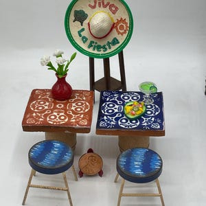 May include: Two miniature tables with blue and red tile tops featuring skull designs. Each table has a blue and white patterned stool. A sombrero with the text "Viva La Fiesta" is on a stand behind the tables.