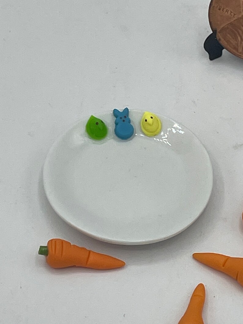May include: A white ceramic plate with three colorful candies: a green leaf, a blue bunny, and a yellow chick. Several orange carrot-shaped candies are scattered around the plate, suggesting an Easter theme.