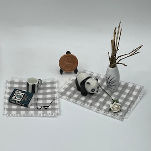 May include: A miniature scene with a black and white panda figurine, a white vase with dried branches, a book titled "Salems Lot" by Stephen King, a striped mug, and a pair of black glasses on a gray and white checkered surface.