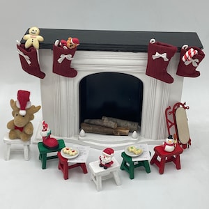 May include: A miniature white fireplace with a black mantel and a black firebox. There are four red stockings hanging from the mantel, each with a different Christmas-themed decoration. There are also several miniature Christmas-themed figurines and furniture pieces around the fireplace, including a reindeer, Santa Claus, a snowman, and a sled.
