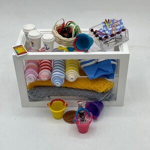 May include: A miniature white display case filled with beach-themed items. Includes coffee cups, a basket of accessories, sunglasses, rolled socks, folded shorts, blankets, and small buckets. A bottle of soda in a pink bucket is also visible.