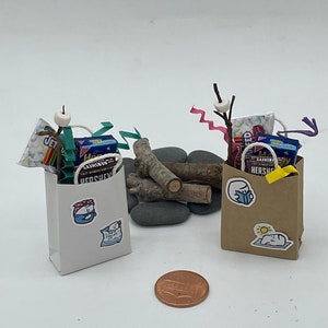 May include: Two miniature camping scenes featuring brown paper bags filled with miniature camping supplies, such as marshmallows, chocolate bars, and graham crackers. The bags are decorated with stickers of a marshmallow wearing a bandana and a marshmallow sunbathing. The scene includes miniature logs, rocks, and a penny.