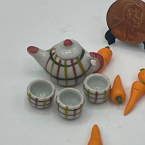 May include: Miniature ceramic tea set with a teapot, two cups, and carrots. The teapot and cups have a white base with a plaid pattern in red, yellow, and green. The carrots are orange with green stems.