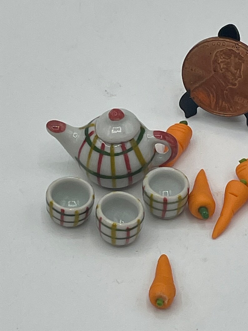 May include: Miniature ceramic tea set with a teapot, two cups, and carrots. The teapot and cups have a white base with a plaid pattern in red, yellow, and green. The carrots are orange with green stems.