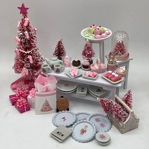 Op de afbeelding: Een miniatuur kersttafereel met een roze flessenborstelboom, zuurstokversieringen en een kleine witte tafel vol miniatuur desserts en decoratieve borden. De scène omvat een miniatuur kerstboom in een glazen koepel en een kleine geschenkdoos.