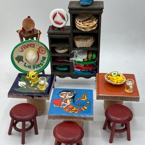 Könnte beinhalten: Ein Miniatur-Restaurantset im mexikanischen Stil mit vier Tischen, vier Hockern, einem Regal mit verschiedenen Gegenständen und einem Zucker Schädel. Die Tische sind mit bunten Fliesen verziert, die eine Meerjungfrau, Blumen und ein traditionelles mexikanisches Design zeigen. Die Hocker sind rot und das Regal ist aus braunem Holz.