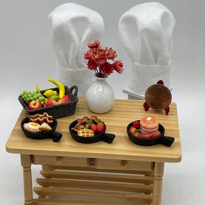 May include: Miniature wooden table with a basket of fruit, small pans of food, a vase of red flowers, and two chef hats. The pans contain waffles, pancakes, and eggs. A penny is on a small stand.
